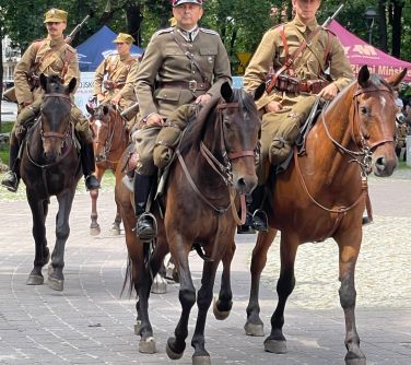 Park, dzień. Aleja z kostki. Ułani w mundurach jada na koniach