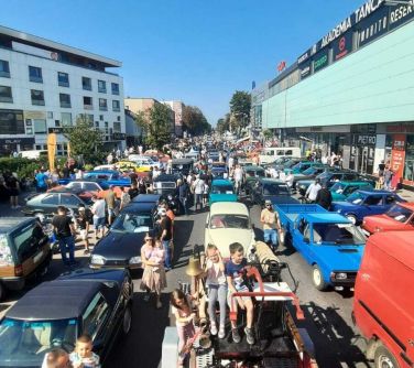 Dwór. Na ulicy i parkingach stoją zabytkowe auta. Pomiędzy nimi osoby. Na przodzie na starym wozie siedzą dzieci. Naokoło...