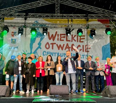 Na dworze. Grupa osób stoi na scenie. Na przodzie przemawia mężczyzna, trzymając rękę z dokumentami w górze