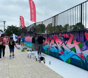 Na dworze. po chodniku przechadzają się osoby. Po prawej mężczyźni maluja na ściance kolorowe grafitti