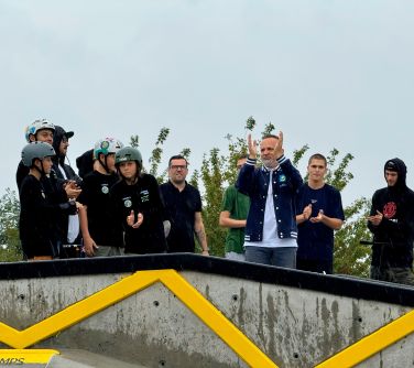 Dwór. Grupa osób w ciemnych koszulkach i kaskach bije brawo. Na przodzie uśmiechnięty mężczyzna klaszcze w dłonie uniesione...