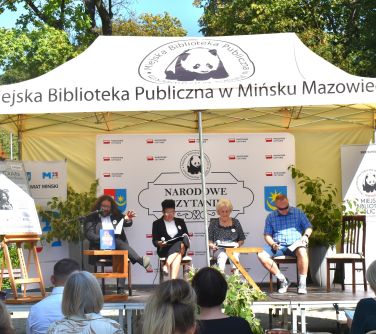Dwór. Cztery sosoby siedzą na krzesłach pod jasnym namiotem czytają książkę