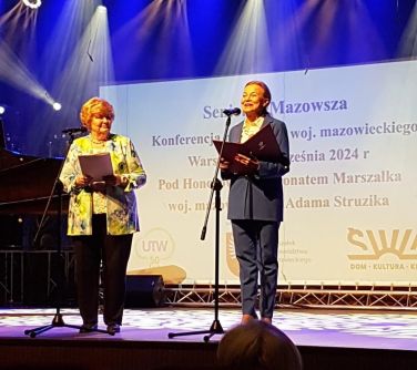 Na oświetlonej scenie dwie kobiety. Jedna z nich przemawia do mikrofonu