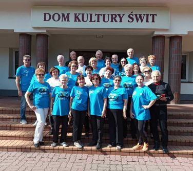 Dwór. Przed jasnym budynkiem z czterema filarami, na schodach pozują seniorzy w niebieskich i białych koszulkach