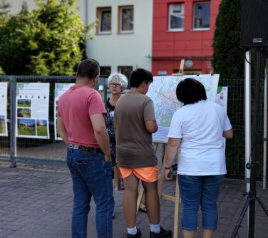 Na zewnątrz. Grupa ludzi stoi przy tablicy z mapą. Rozmawiają.