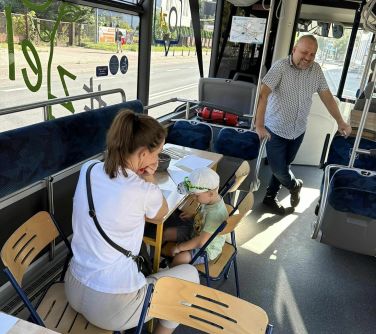 Wnętrze autobusu. Dziecko koloruje, obok siedzi kobieta. W tle stoi uśmiechnięty mężczyzna.