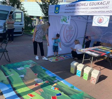 Na zewnątrz. Na chodniku leży kolorowa gra dla dzieci, obok namiot. W jego środku są ludzie.