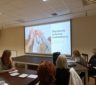 Sala. Na przodzie duży ekran z treścią. Przy stole dwie kobiety. Jedna czyta. Trzy osoby obserwują i słuchają