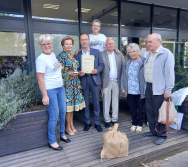 Na zewnątrz. Ośmiu seniorów pozuje do zdjęcia przed oszklonym budynkiem. Naokoło zielone krzewy