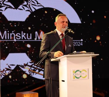 Pomieszczenie. Mężczyzna w ciemnym garniturze przemawia sojąc przed jasną mównicą