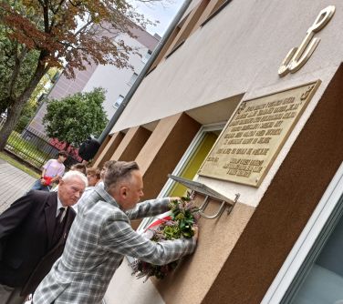 Na zewnątrz. Mężczyzna w marynarce w szarą kratę wiesza wianuszek z kwiatów na ścianie. Obok stoi drugi mężczyzna