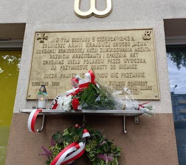 Na zewnątrz. Na ścianie budynku pod wiszącą tablicą i kotwicą jest półka ze zniczem i kwiatami. Pod nimi wisi wianuszek