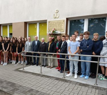 Grupa osób stoi pod budynkiem szkoły przy podjeździe