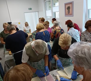 Pomieszczenie. Grupa seniorów w rękawiczkach i faruchach stoją przy stołach i gotują