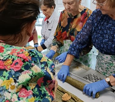 Pomieszczenie. Cztery seniorki w fartuchach i niebieskich rękawiczkach stoją przy stole i gotują