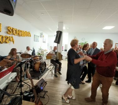 Pomieszczenie. Seniorzy tańczą. Po lewej gra orkiestra. Na jasnej ścianie z balonów ułożony napis dzień seniora 2024
