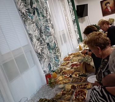 Pomieszczenie. Przy jasnej ścianie stoją stoły wypełnione jedzeniem. Obok nich seniorzy