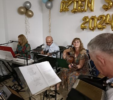 Pomieszczenie. Zdjęcie siedzącej i grającej orkiestry. Nad nimi na jasnej ścianie złote balony dzień seniora 2024