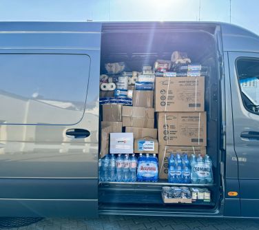 Na zewnątrz. Bok auta transportowego z otwartymi bocznymi drzwiaki. Samochód wypełniony pudłami