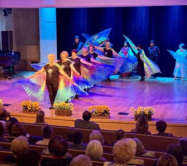 Sala koncertowa. Na scenie występują seniorzy w czarnych ubraniach i przyczepionymi dużymi świecącymi skrzydłami motyli. Na...