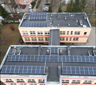 Dwór. Dachy dwóch budynków z instalacją fotowoltaiczną. Po lewej sucha trawa. Na przodzie budynki, drzewa i auta na parkingu
