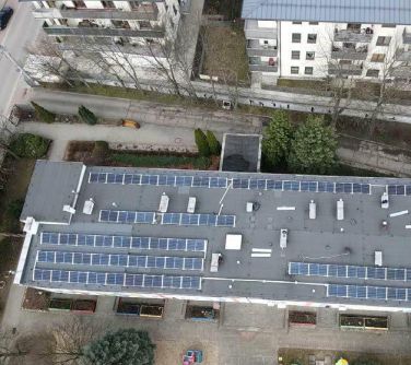 Dwór. Dach budynku z instalacją fotowoltaiczną. Naokoło trawy, chodnik i drzewa. Na przodzie dwa budynki