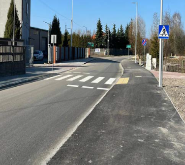 Na zewnątrz, słonecznie. Ulica z przejściem dla pieszych, po prawej i lewej chodnik. Po lewej budynki