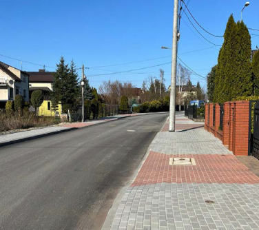 Na zewnątrz, słonecznie. Ulica, po lewej i prawej chodnik z kostki. Po prawej drzewa na posesji. Po lewej budynki
