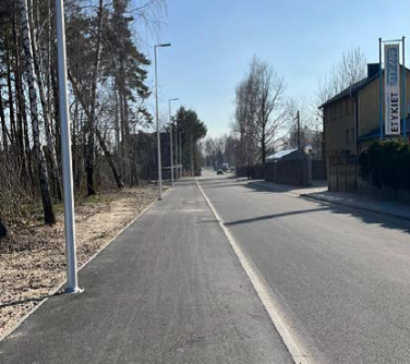 Na zewnątrz, słonecznie. Ulica, po lewej gładki chodnik, latarnie i drzewa. Po prawej budynki