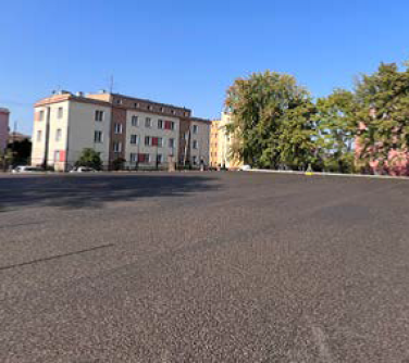 Na dworze, słonecznie. Pusta nawierzchnia boiska. W tle budynki i drzewa. Po prawej wysokie, zielone ogrodzenie