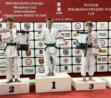 Pomieszczenie. Na podium na trzech miejscach stoją chłopcy w białych kimonach. trzymaja nagrody i dyplomy. W tle duży baner...