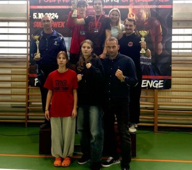 Hala sportowa, dzień. Osiem osób stoi przed banerem zawieszonym na drewnianych drabinkach. Trzy osoby trzymają puchary