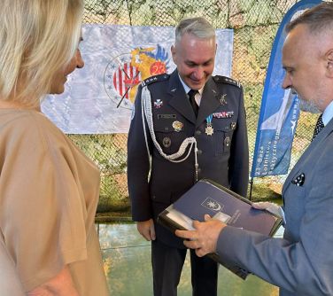 Pomieszczenie. Kobieta i mężczyzna wręczają list gratulacyjny dowódcy z mundurze.