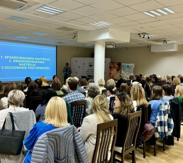 Sala konferencyjna. Na krzesłach siedzą osoby. Na przodzie mężczyzna opowiada stojąc przy dużym niebieksim ekranie