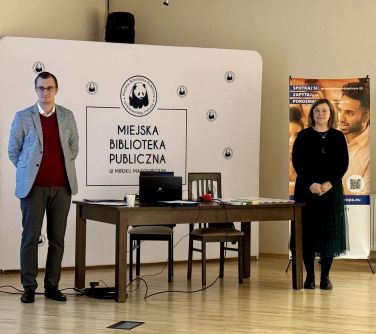 Sala konferencyjna. Stół, po lewej mężczyzna, po prawej kobieta. W tle duży, biały baner. Na stole laptop i dokumenty