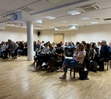 Sala konferencyjna. Na krzesłach siedzą osoby. Po środku biała kolumna