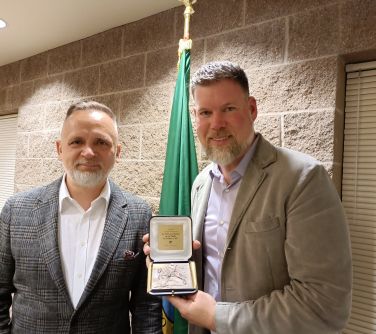 Pomieszczenie. Dwóch, usmiechniętych mężczyzn. Po prawej trzyma otwarty medal z dedykacją. W tle z grubych cegieł ściana i...
