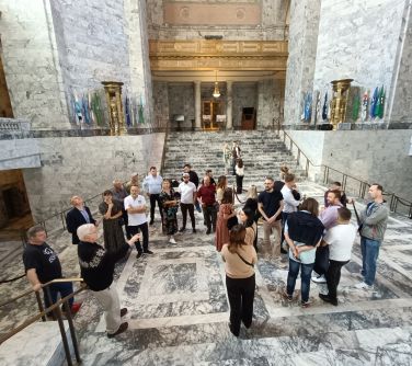 Pomieszczenie ze schodami i w marmurze. Grupa osób ogląda, słucha