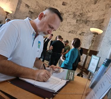 Pomieszczenie. Mężczyzna w białej koszulce wpisuje się do dziennika leżącego na drewnianym podwyższeniu. W tle osoby i...