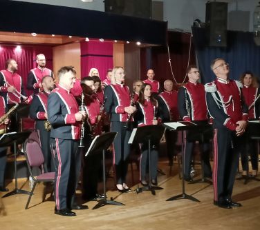 Pomieszczenie. Orkiestra w galowych mundurach czrno-czerwonych z instrumentami w rękach stoją przed krzesłami. Na ich...