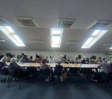 Pomieszczenie, dzień. Trzy podłużne okna w suficie. Przy długim stole na krzesłach siedzą osoby