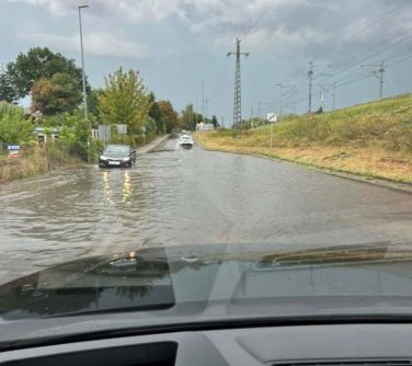 Na dworze, pochmurny dzień. Zdjęcie przez szybę auta na zalaną ulicę. Na przodzie po lewej auto przejedża przez wodę...
