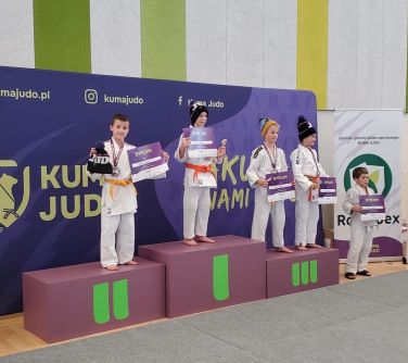 Pomieszczenie. Na wrzosowym podium czterech chłopców. Dwóch na trzecim miejscu. Po prawej piąty chłopiec. W tle banery