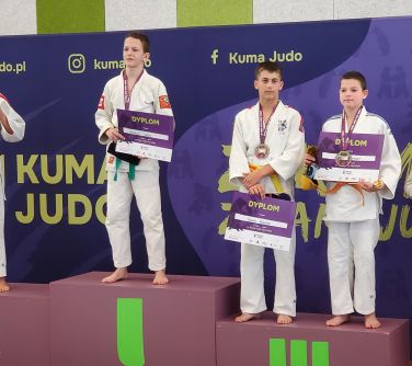 Pomieszczenie. Czterech chłopców w białych kimonach na podium. Dwóch na miejscu trzecim. Po prawej piąty chłopiec. Po obu...
