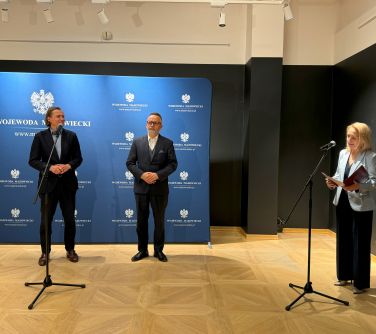 Pomieszczenie. Dwa stojące mikrofony. Po prawej przemawia kobieta, czyta z kartki. Po lewej dwóch mężczyzn. Jeden stoi...