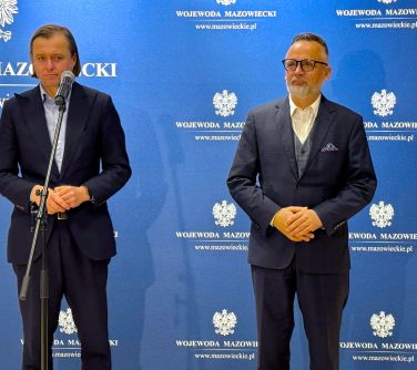 Dwóch mężczyzn w ciemnych garniturach. W tle niebieska ścianka. Po lewej mikrofon stojący