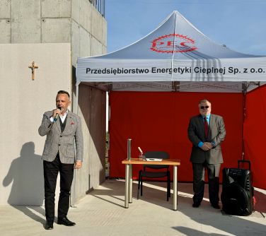 Na dworze, słonecznie. Dwóch mężczyzn. Jeden przemawia do mikrofonu. W tle namiot szaro-czerwony