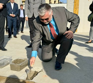 Na dworze, słonecznie. Mężczyzna kuca. Kielnią nabiera cement z plastikowego pojemnika