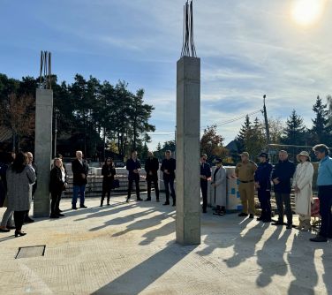 Na dworze, słonecznie. Na placu w półkolu grupa ludzi. Pomiędzy dwie betonowe, kwadratowe kolumny zakończone metalowymi...