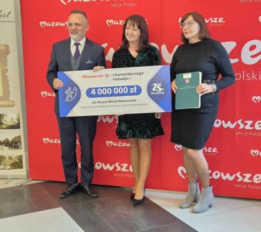 Pomieszczenie. na tle dużego, czerwonego baneru trzy osoby pozują, trzymając duże kartonowe czeki